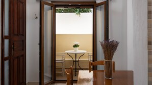 Ein Zimmer mit Holztür, einem kleinen Tisch mit weißer Vase und Blick auf einen Balkon mit weißer Wand und einer Pflanze.