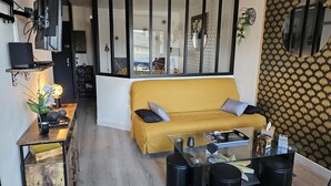 Ein gemütlicher Wohnbereich mit gelbem Sofa, einem Glastisch, schwarzen Poufs, einem an einer Holzablage montierten Fernseher und einer Wand mit Muster.