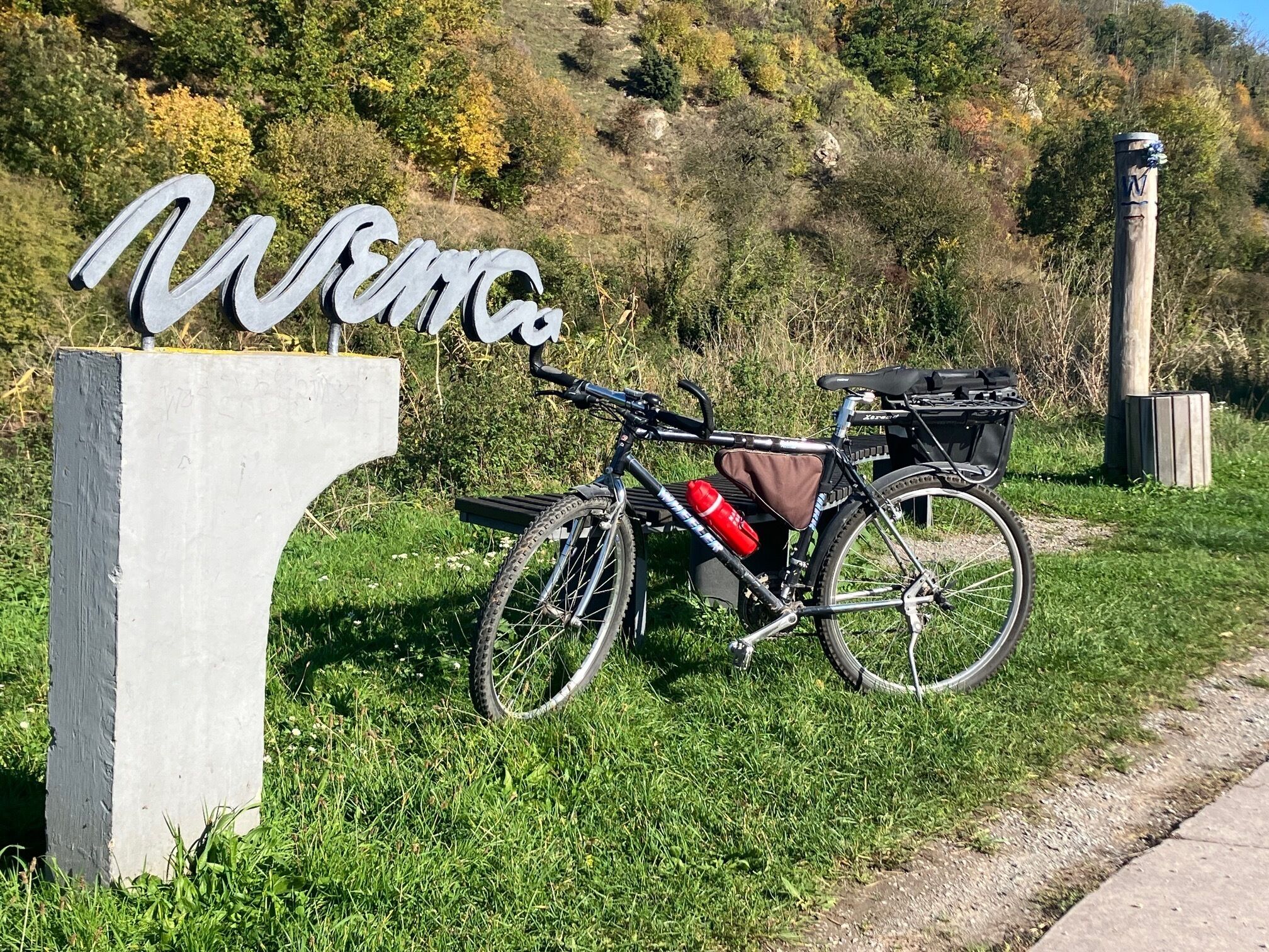 A bicycle parked next to a sign with the word "Welcome" on a grassy area with a hilly background.