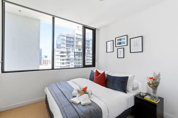 A modern bedroom with a large window, a neatly made bed, and a nightstand with a lamp and books.