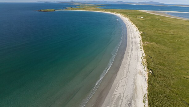 An aerial view of a coastal landscape with a curved beach, grassy areas, and distant mountains. - Heisgeir (Isle of North Uist)