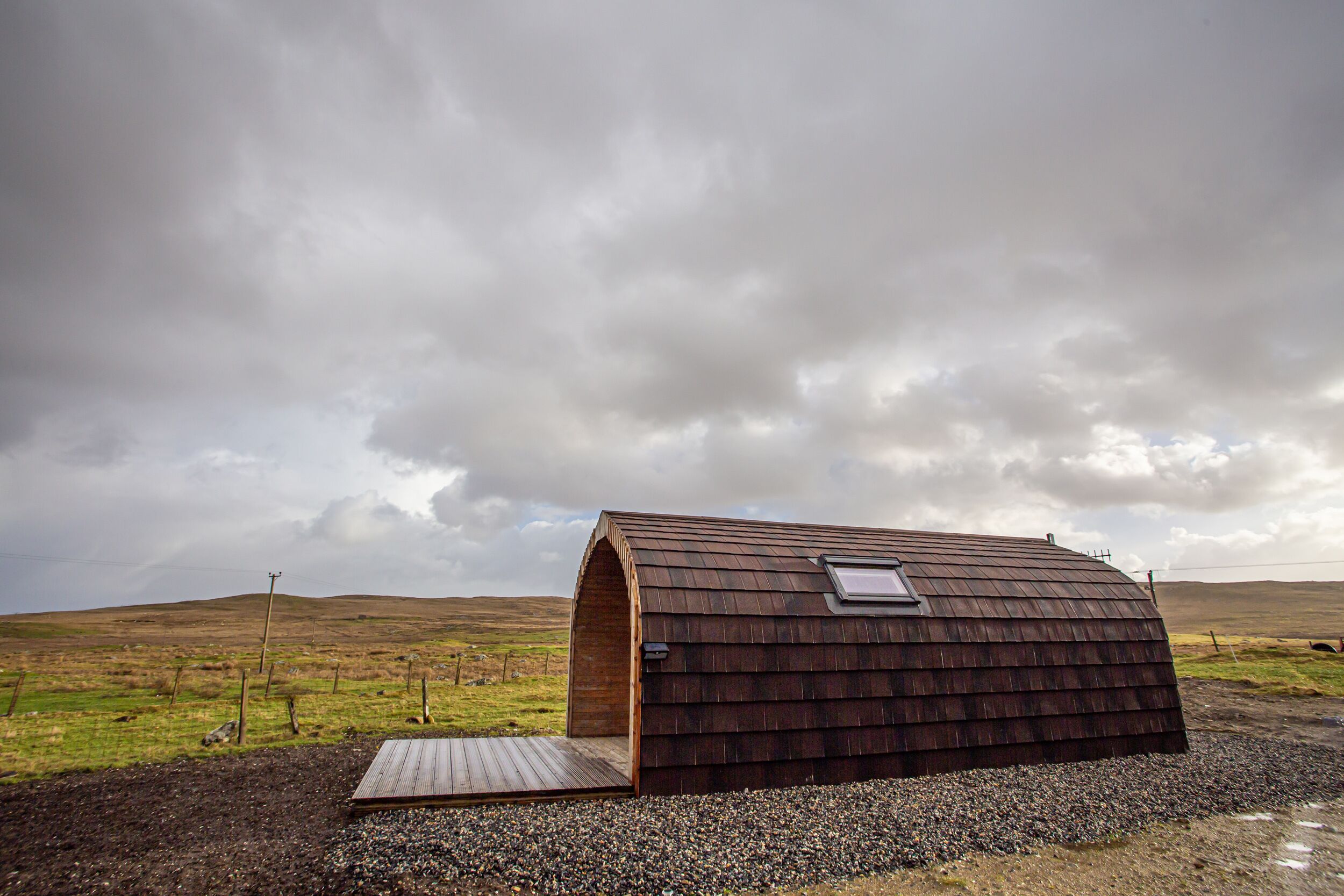 A single-story, wooden structure with a sloped roof, situated in a rural landscape with a gravel path leading to it.