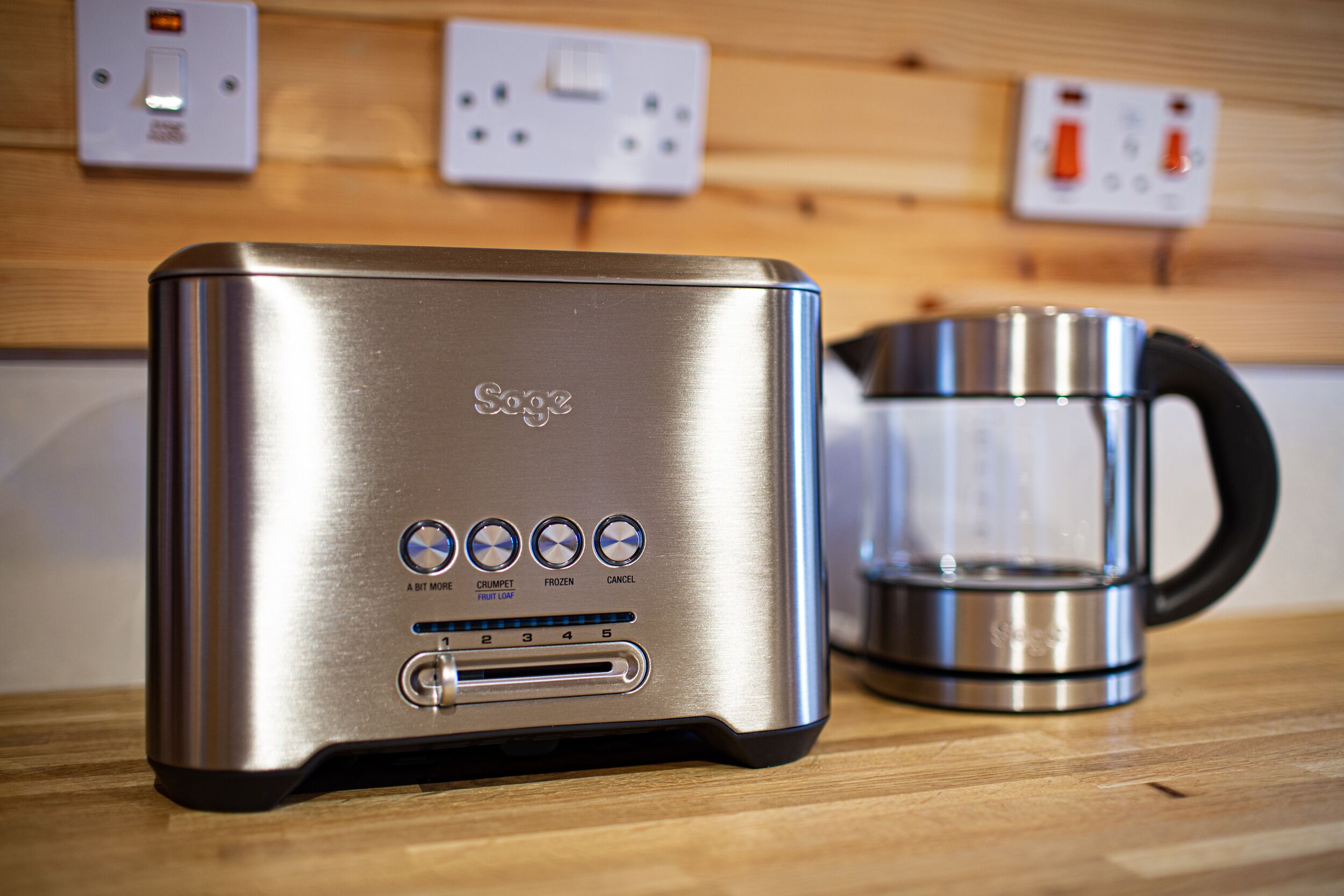 A stainless steel toaster with a digital display and multiple settings, placed on a wooden surface with a coffee maker nearby.
