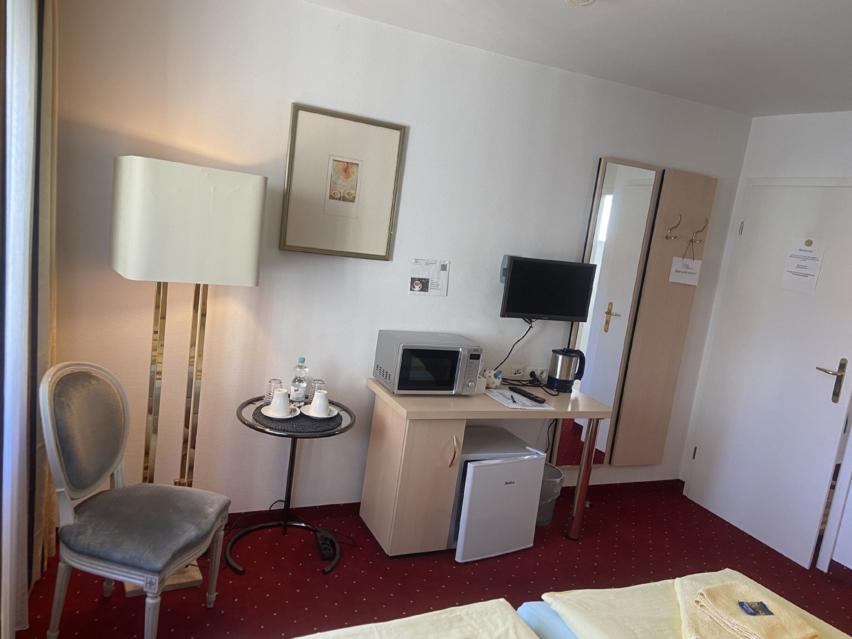 A hotel room with a desk, a chair, a bed, a small refrigerator, a microwave, a television, and a bedside table with a lamp.