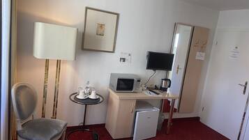 A hotel room with a desk, a chair, a bed, a small refrigerator, a microwave, a television, and a bedside table with a lamp.