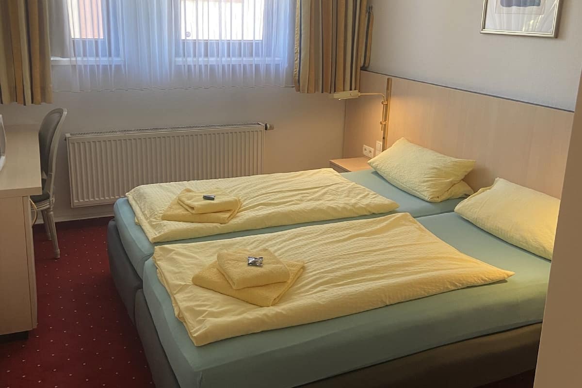 A hotel room with two beds, a desk, a chair, a radiator, a lamp, and a window with curtains.