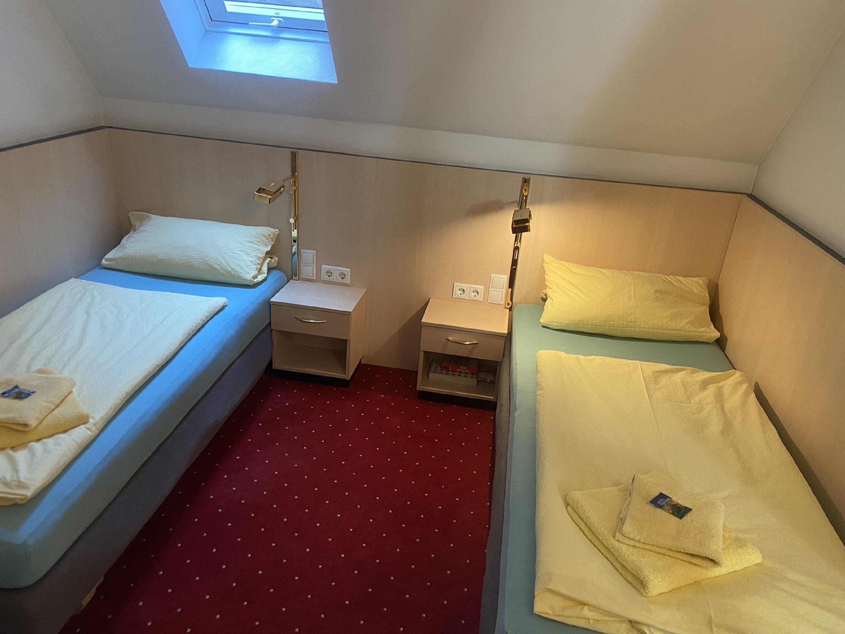 A small, compact room with two single beds, each with a pillow and a folded towel. There is a small bedside table with a lamp on one side and a power outlet on the other. The room has a red carpet with white dots and a skylight above the beds.