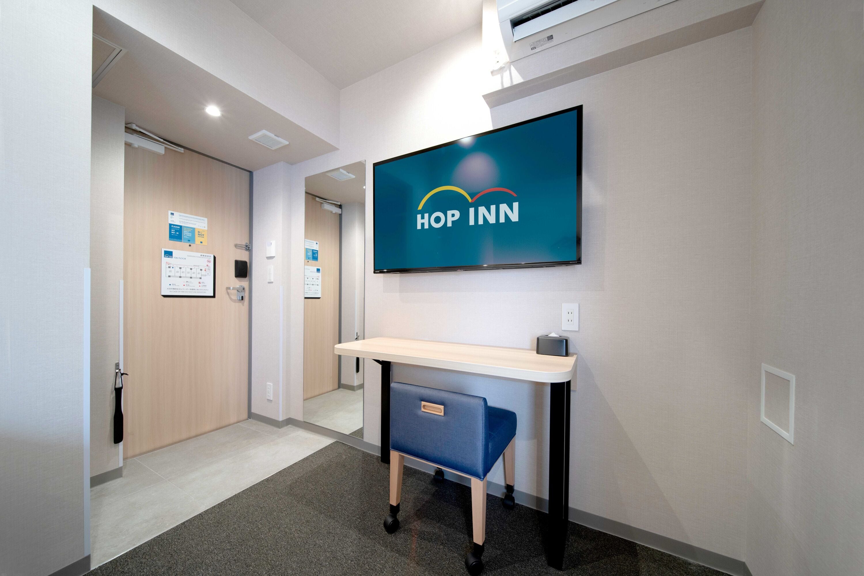 A hotel reception area with a reception desk, a mounted television displaying the 'Hop Inn' logo, and a wall-mounted air conditioning unit.