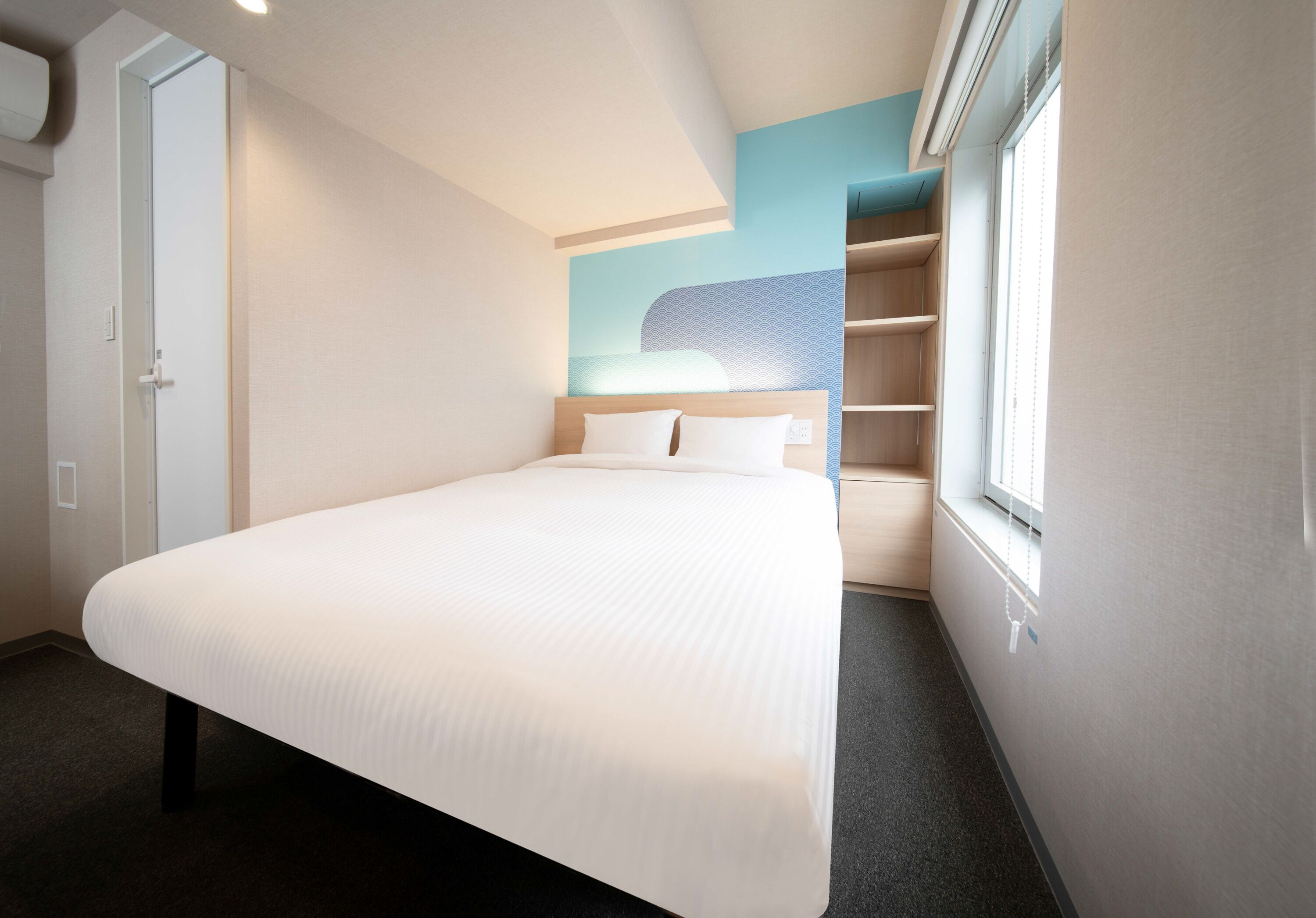A hotel room with a large bed, a wardrobe, a built-in shelf unit, and a window with blinds.