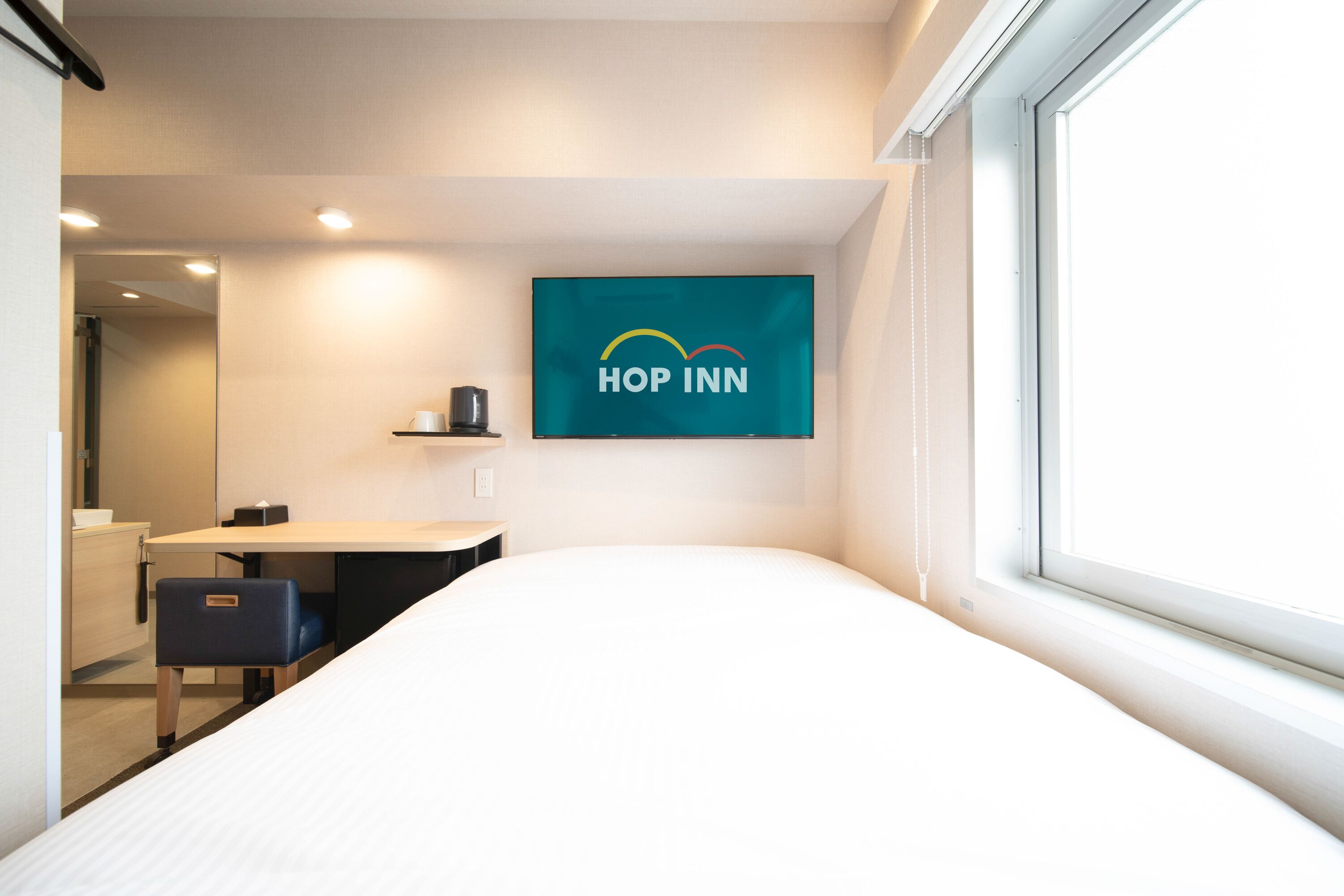A modern hotel room with a bed, a desk, a chair, a small shelf with a cup, and a window.