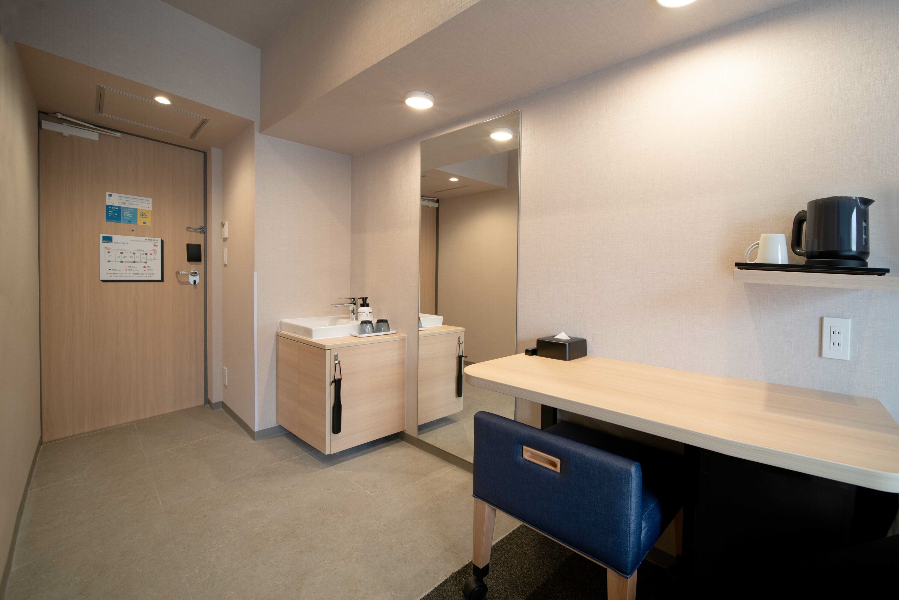 Une chambre d’hôtel moderne avec une porte en bois, un lavabo avec un miroir, une petite table avec une chaise et une étagère avec une bouilloire et une tasse.