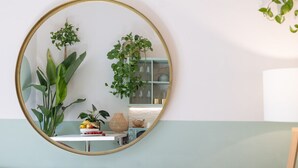 Un espejo redondo con marco dorado que refleja una habitación con pared azul claro, una mesa blanca con un frutero y una planta.