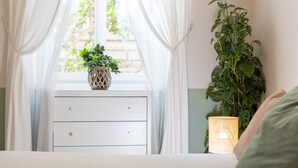 Un dormitorio con ventana, una mesita de noche blanca con cajones, una planta en maceta y una lámpara encendida.