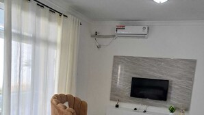Un salon moderne avec un canapé beige, un fauteuil marron, un téléviseur à écran plat fixé à un mur gris et un appareil de climatisation blanc.