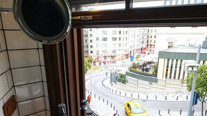 A bathroom with a white sink, a mirror, and a window overlooking a city street with buildings and a yellow taxi. - Hostel Le Banc (Istanbul)