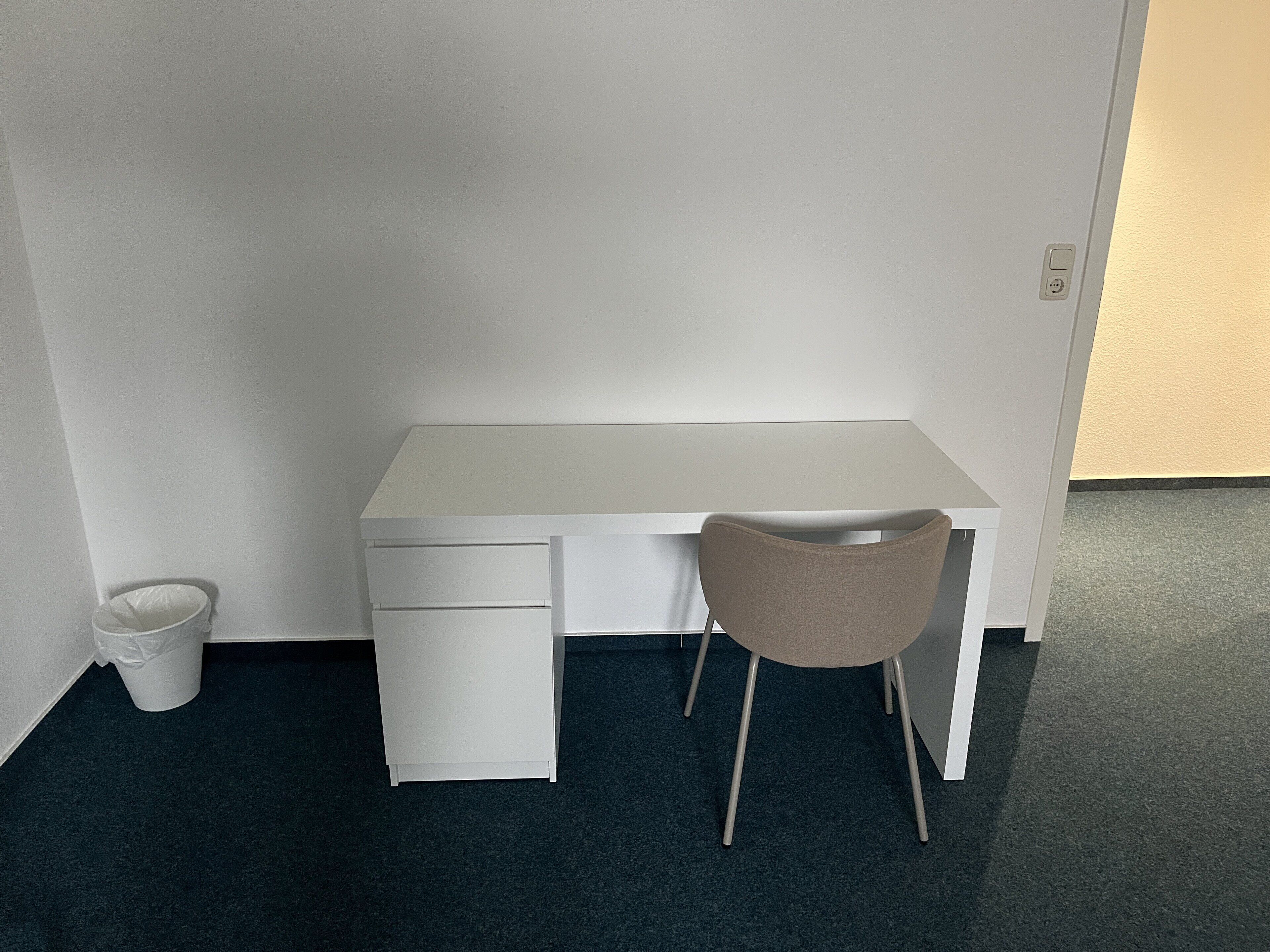 Een wit bureau met een stoel, een prullenbak en een deur.