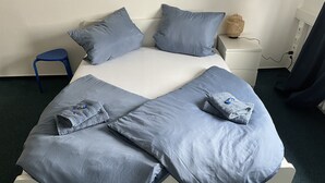 Ein ordentlich bezogenes Bett grau, Kissen, ein kleiner Nachttisch mit Lampe und ein Sessel.