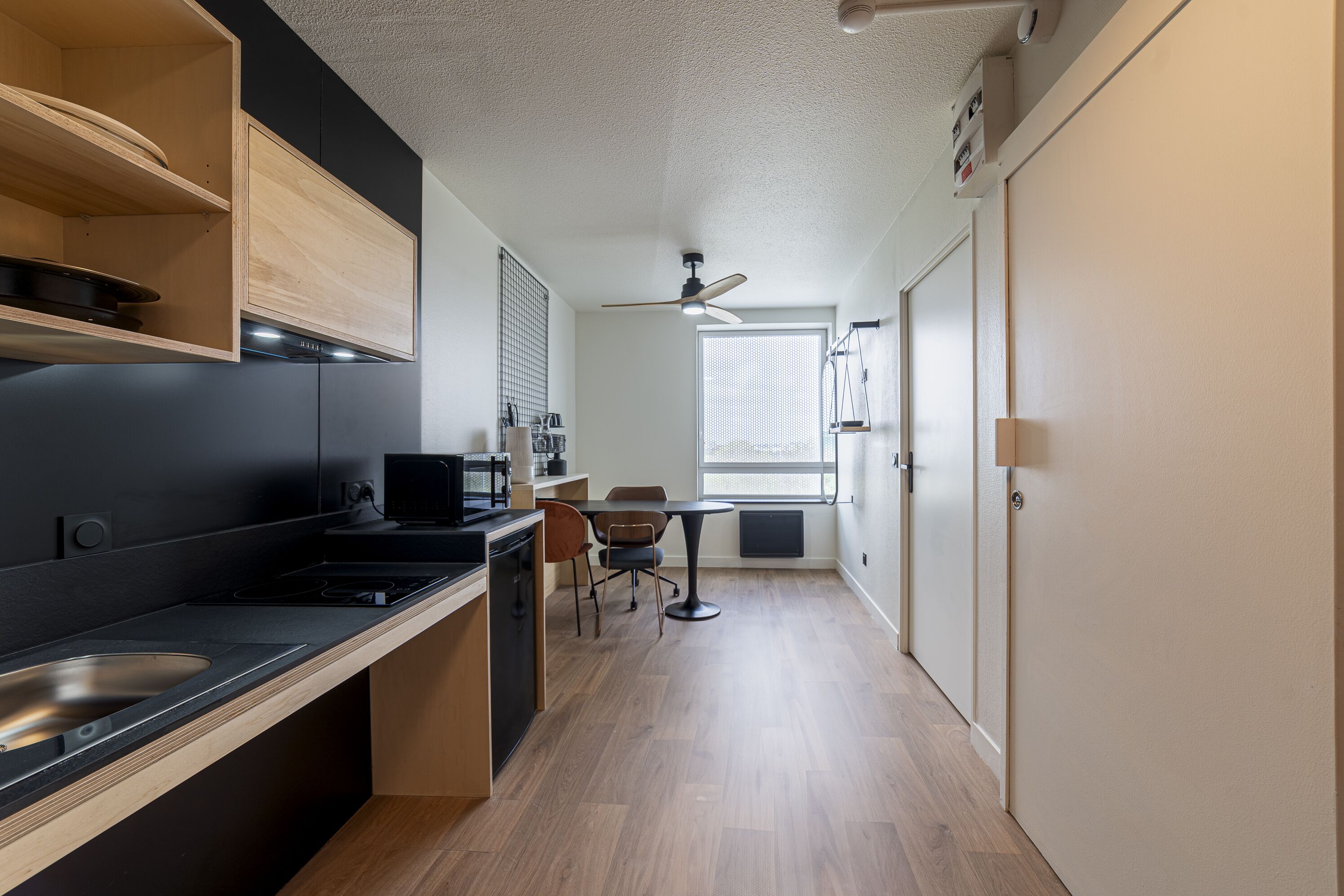 A compact kitchen with a microwave, stove, and sink. There is a dining area with a table and chairs. A ceiling fan is visible, and a window provides natural light.