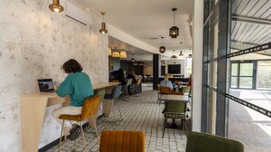 A modern cafe with a rustic wall, wooden tables, and chairs, and a large window offering a view of the outdoors. - Cowool Toulouse (Toulouse)