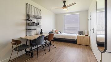 A compact bedroom with a bed, a desk with chairs, a ceiling fan, and a window with blinds.