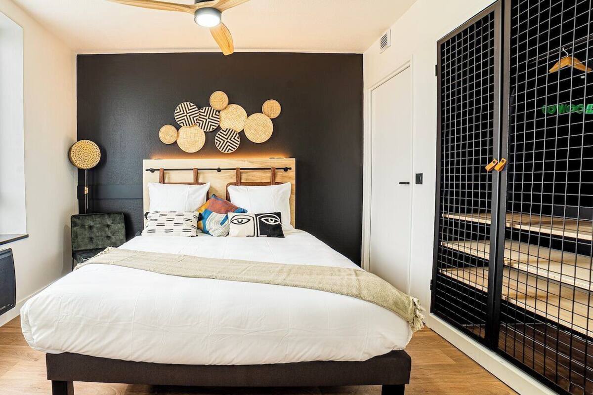 Une chambre moderne avec un grand lit, un ventilateur de plafond, un élément décoratif mural et une vue sur un balcon agrémenté d’une grille en métal noir.