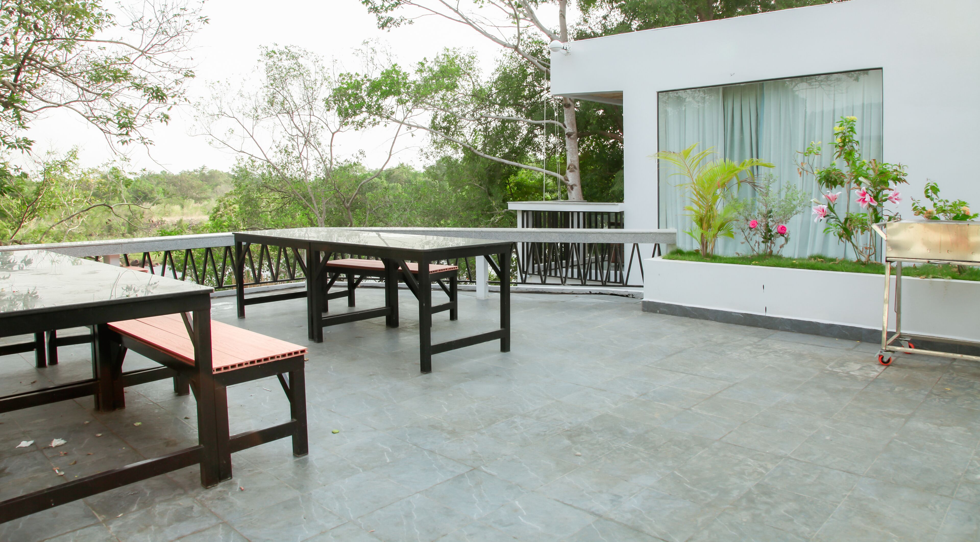 An outdoor patio with tables and chairs, a modern building with large windows, and a garden area with plants and flowers.