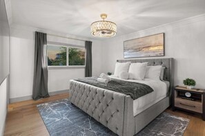 A bedroom with a tufted bed, a chandelier, a painting, a nightstand, and a window with curtains.