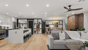 Une cuisine moderne avec un îlot central, un coin repas agrémenté d’un mur en briques, et un salon avec un canapé et un ventilateur de plafond.