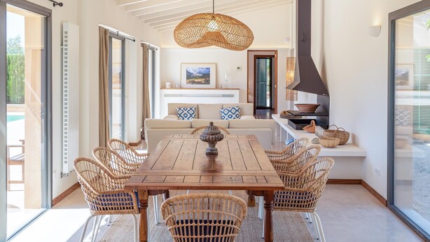 Uma sala de jantar com uma mesa de madeira e cadeiras de vime, um lustre e vista para o exterior.