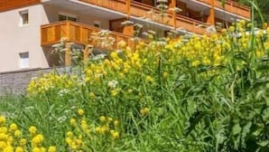 A wooden chalet with multiple balconies, surrounded by lush greenery and yellow flowers. - T2 Montagne Cosi Pied des Pistes- Piscine Hammam (Allos)