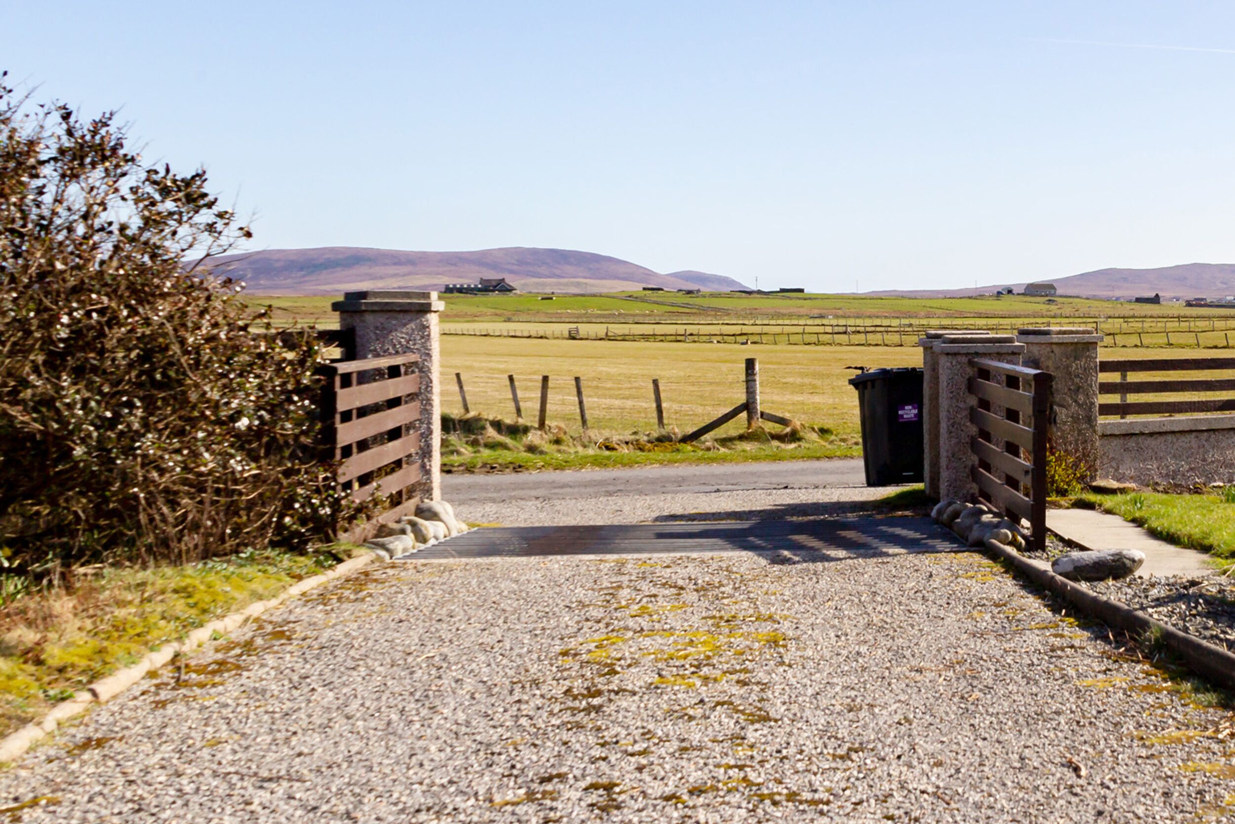 A rural road leading to a gated property with a view of distant hills.