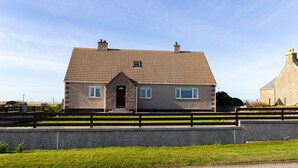 A two-story house with a tiled roof, a front porch, and a stone foundation. - Ar Dachaigh (Isle of North Uist)