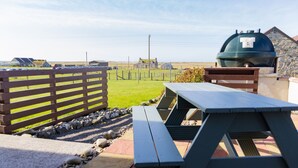 A picnic table and grill area with a view of a grassy field and distant buildings. - Ar Dachaigh (Isle of North Uist)