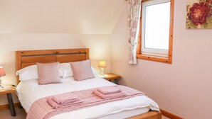 A bedroom with a wooden bed, a nightstand with a lamp, a window with curtains, and a floral painting on the wall. - Ar Dachaigh (Isle of North Uist)