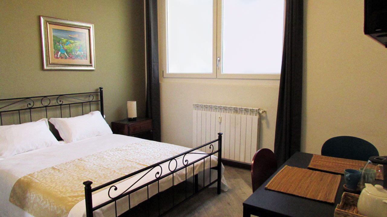 Ein Hotelzimmer mit Bett, Tisch mit Teekanne, einem Bild an der Wand und einem Fenster mit Vorhängen.