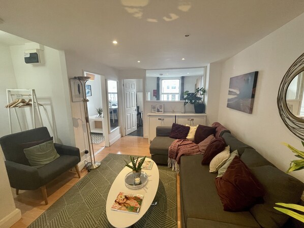 Ein modernes Wohnzimmer mit einer Couch, einem Sessel und einem Couchtisch. Durch einen offenen Durchgang ist ein Essbereich zu sehen.