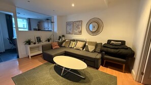 Ein modernes Wohnzimmer mit einem Ecksofa, einem runden Couchtisch und einem kleinen schwarzen Schlafsofa. An den Wänden hängen zwei gerahmte Kunstwerke und über dem Sofa befindet sich ein Spiegel.