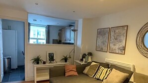 A modern living room with a sofa, a coffee table, and a patterned rug. There is a kitchen area in the background with a window and a plant. - Cosy two bedroom flat Fulham Broadway (London)