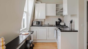 Cocina compacta con armarios blancos, encimera negra, microondas, cafetera y ventana que proporciona luz natural.
