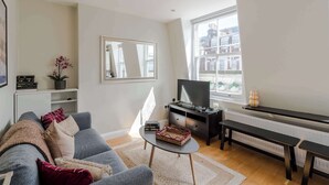 Una sala de estar con un sofá gris, una mesa de centro, un televisor, un piano y un amplio ventanal con vistas a edificios.