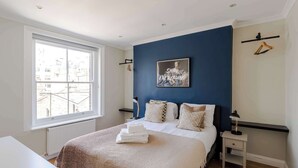 Un dormitorio con un ventanal grande, una cama con ropa de cama blanca, una pared de acento azul y un cuadro enmarcado.