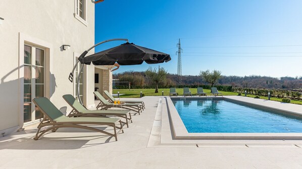 A swimming pool with lounge chairs and an umbrella, a clear sky, and a view of a hill.
