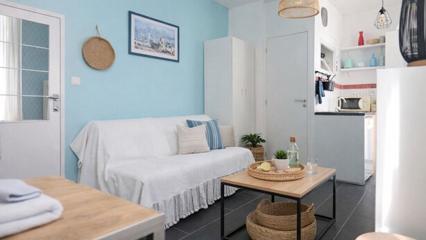 A modern living room with a blue wall, a white sofa, a wooden coffee table, and a kitchen area with a microwave and cabinets. - Seafront studio close to all amenities (Les Sables-d´Olonne)