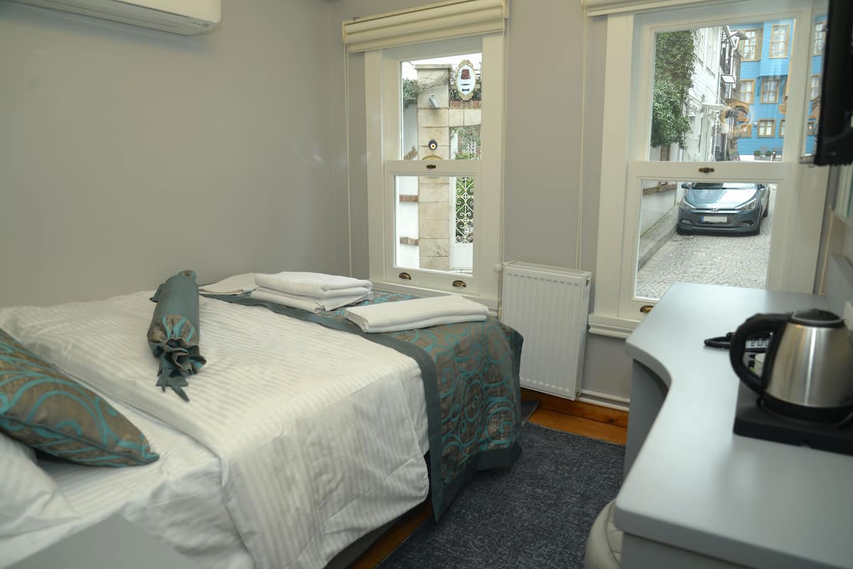 A neatly made bed with a white and teal bedspread, a window with a view of a street and parked car, and a kettle on a counter.