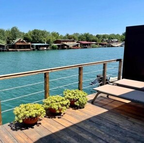 Una terraza de madera con plantas en macetas, barandilla y vistas a casas al otro lado del agua.