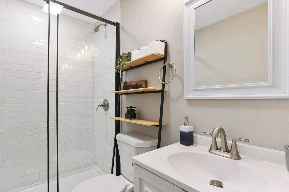 Une salle de bain moderne avec une douche à parois de verre, un lavabo blanc doté d’un robinet doré et une étagère en bois où sont rangés les articles de toilette.