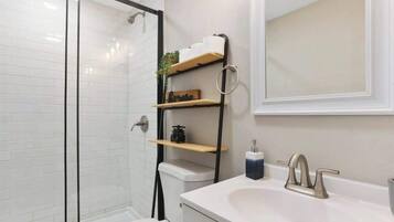 Une salle de bain moderne avec une douche à parois de verre, un lavabo blanc doté d’un robinet doré et une étagère en bois où sont rangés les articles de toilette.