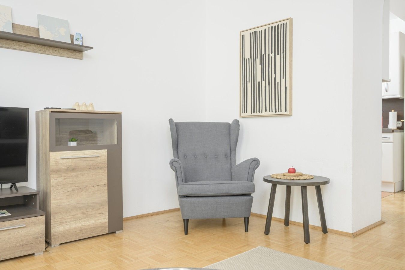 Een moderne woonkamer met een grijze fauteuil, een klein rond tafeltje met een rode appel, en een flatscreen televisie op een houten kast.