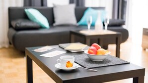 Een moderne woonkamer met een donkere bank, een klein bijzettafeltje met een schaal fruit en een beker, en een eettafel met een placemat.