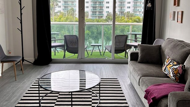 Ein modernes Wohnzimmer mit einem grauen Sofa, einem runden Glastisch und einem schwarz-weiß gestreiften Teppich. Ein Balkon mit Gartenmöbeln bietet Blick auf hohe Gebäude.